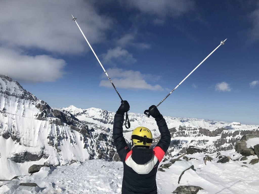 A person with a yellow helmet and black jacket standing on a snowy mountain with ski poles.