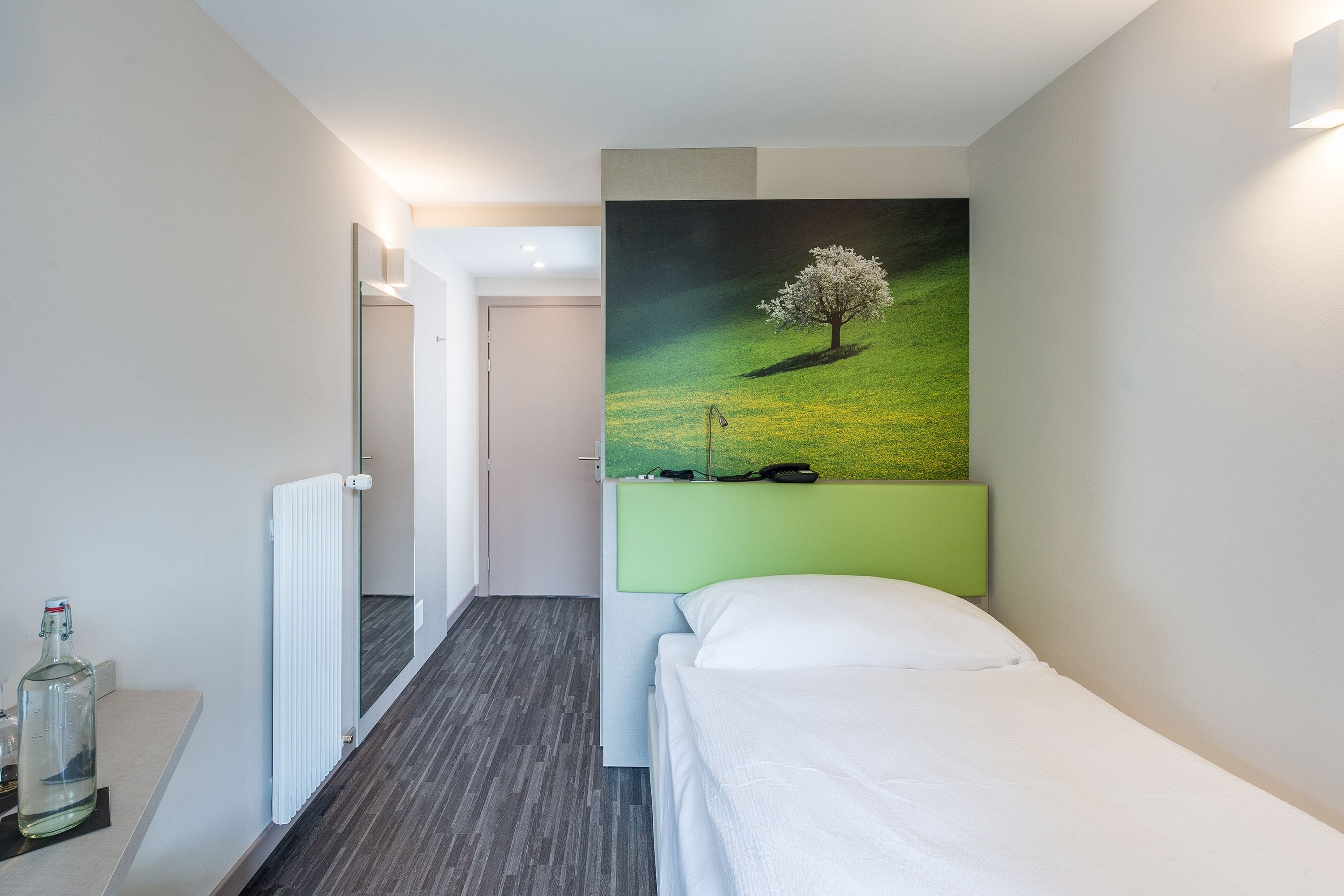 Hotellihuone, jossa on sänky, maalaus, jossa on kuvattuna puu, patteri ja pullo yöpöydällä.