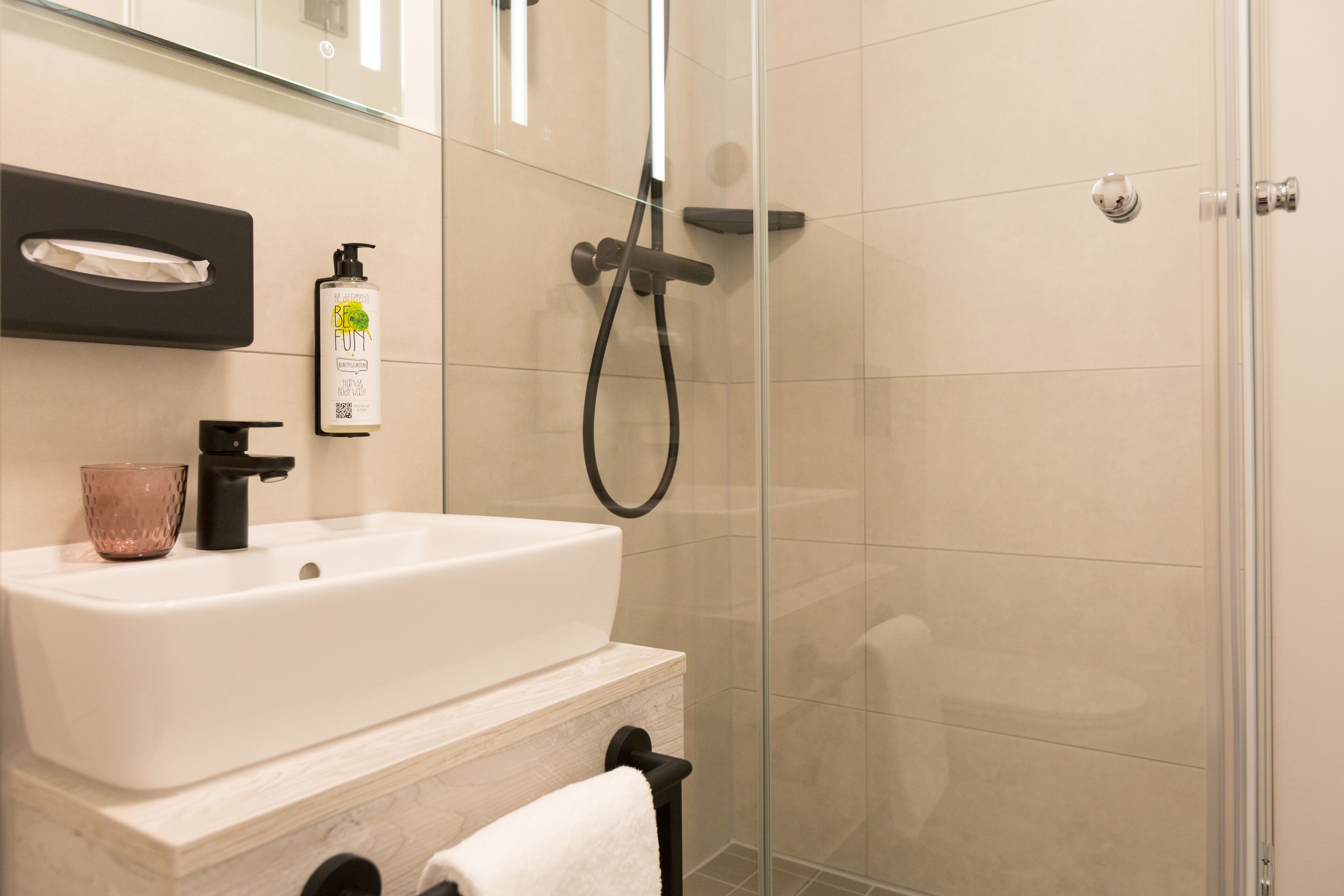 Une salle de bain avec un lavabo blanc, un robinet noir, une cabine de douche en verre et un sèche-mains.