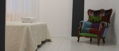 A room with a bed covered in a white bedspread, a chair with colorful cushions, and a small table with folded towels.