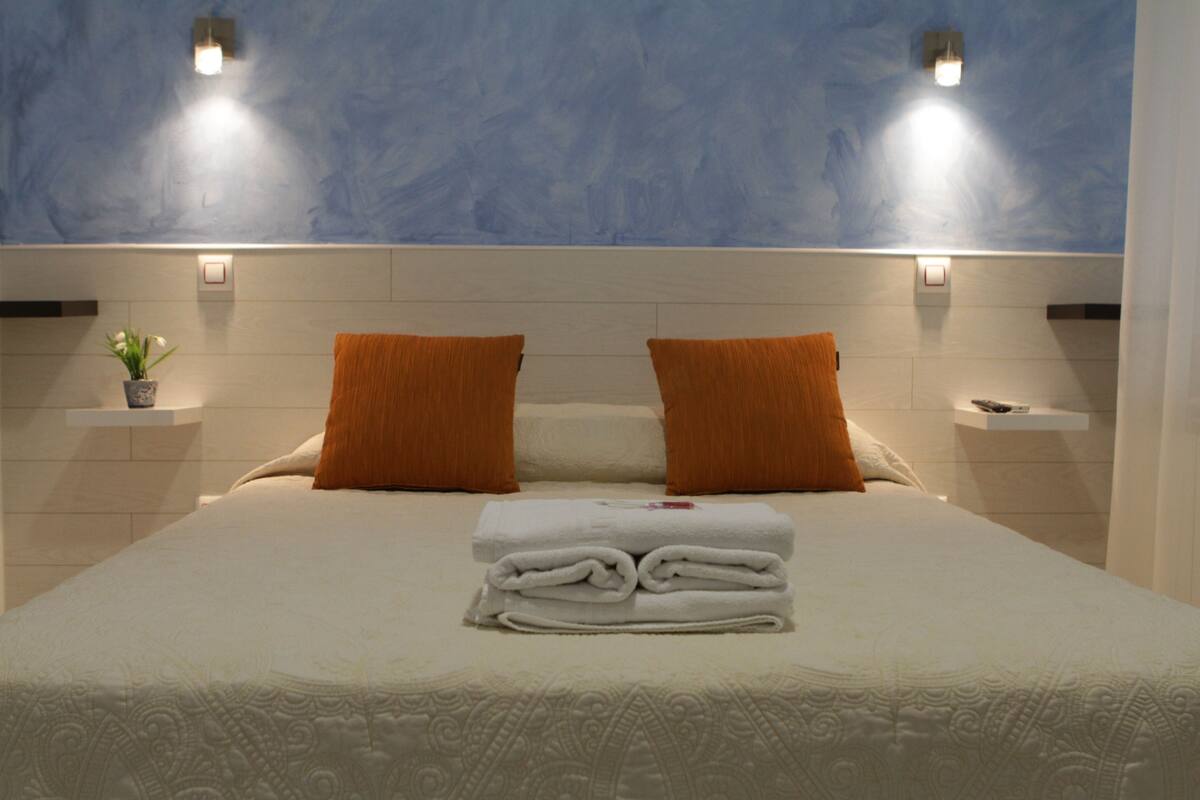 Una cama bien hecha con toallas blancas dobladas, dos almohadas naranjas y una pequeña planta en maceta sobre una mesita de noche.