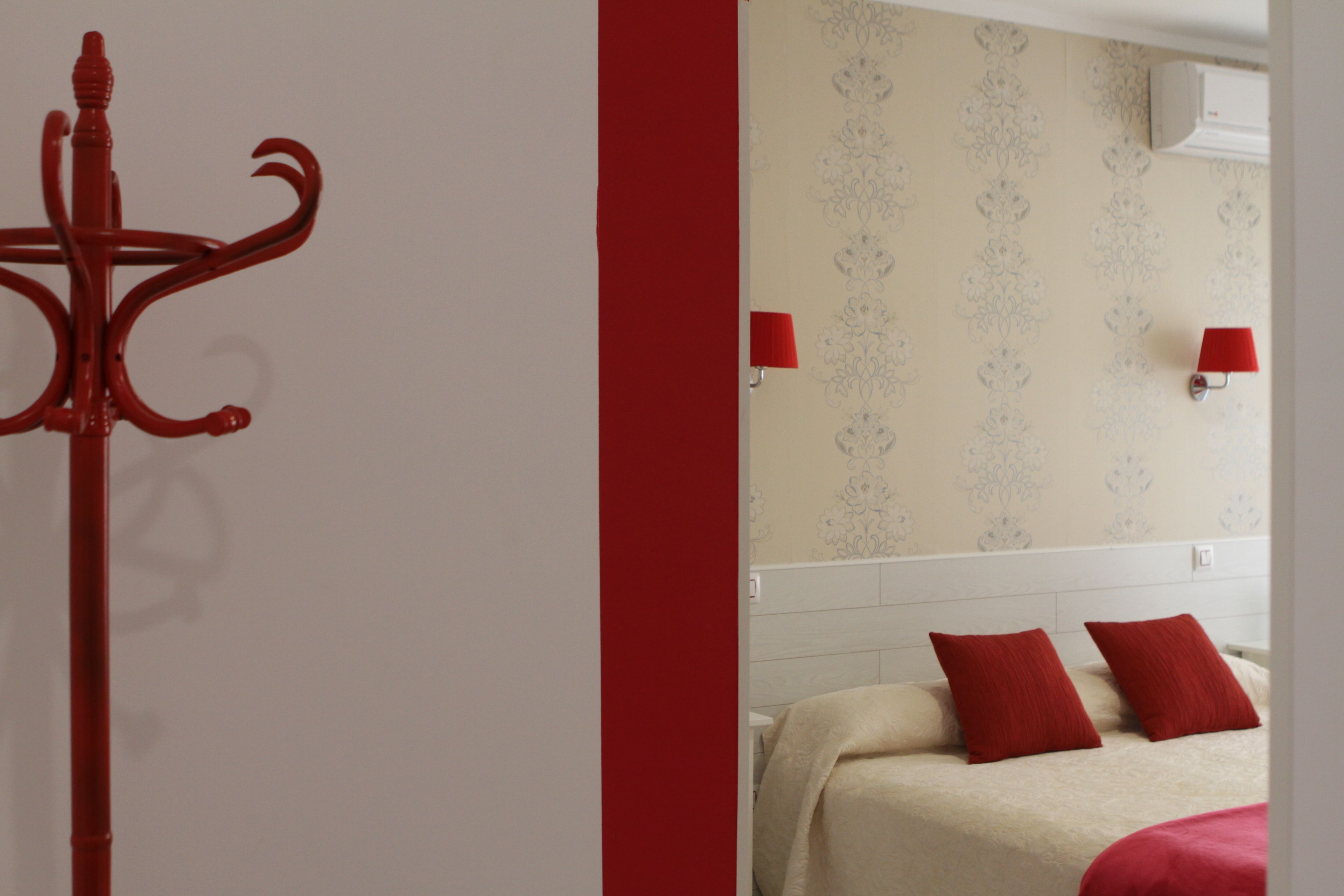 A hotel room with a bed, two red pillows, a red coat rack, and a wall with a floral pattern.