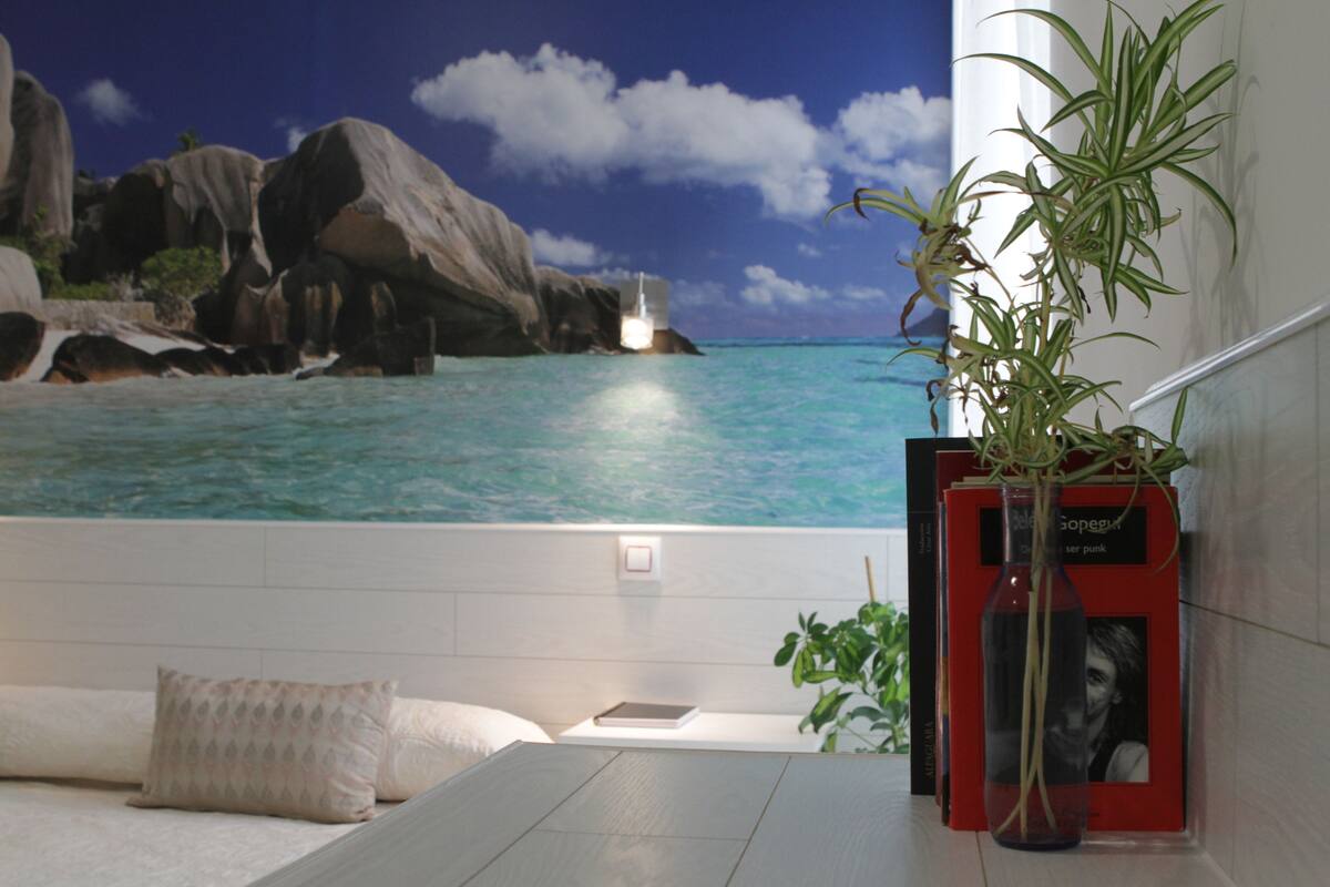 Una habitación con vistas a una costa rocosa y al océano, una cama con almohadas, una mesita y una planta en maceta.