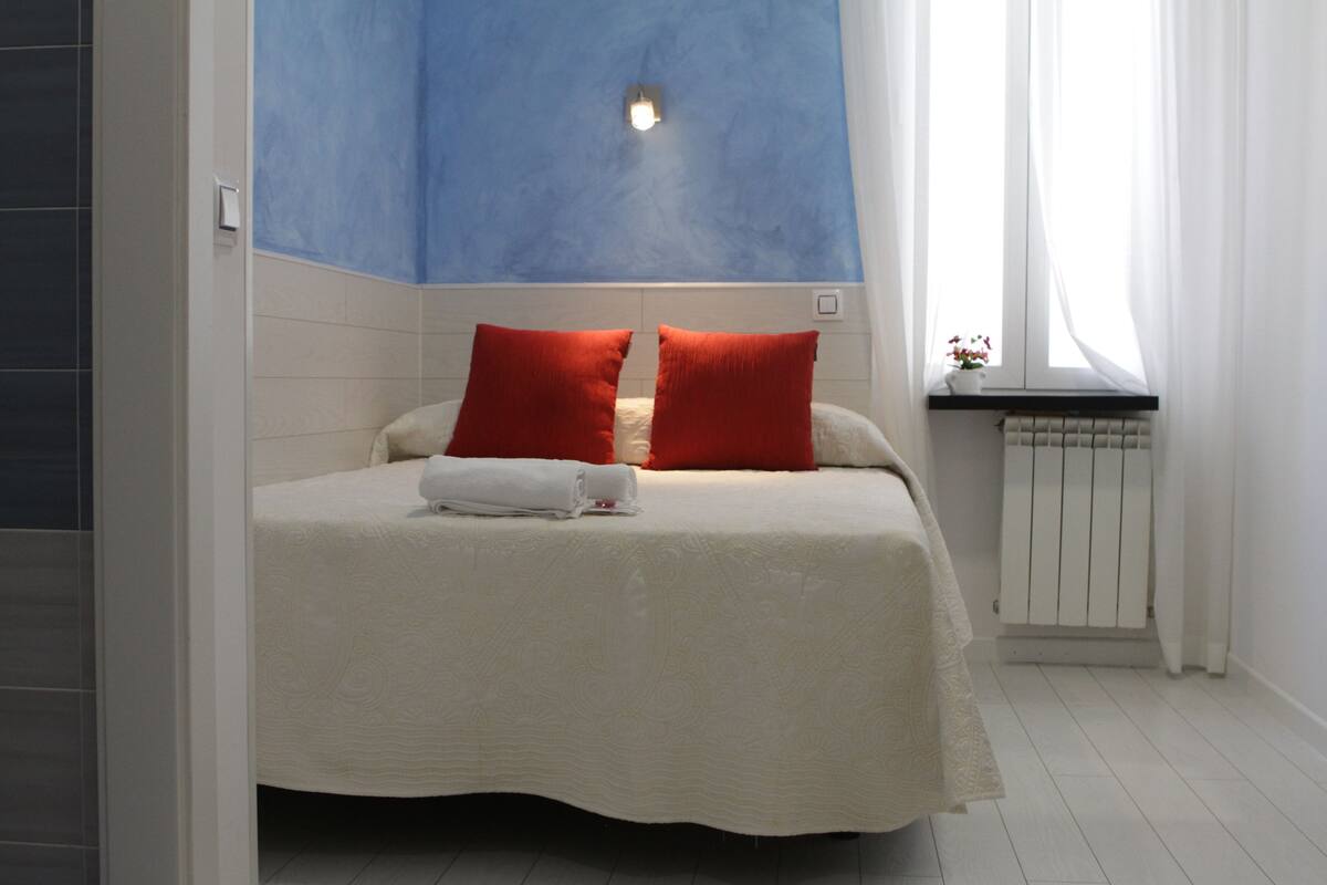 Un dormitorio con cama, dos almohadas rojas, una colcha blanca, una pared azul decorativa, una ventana con vistas y una mesita con un jarrón de flores.