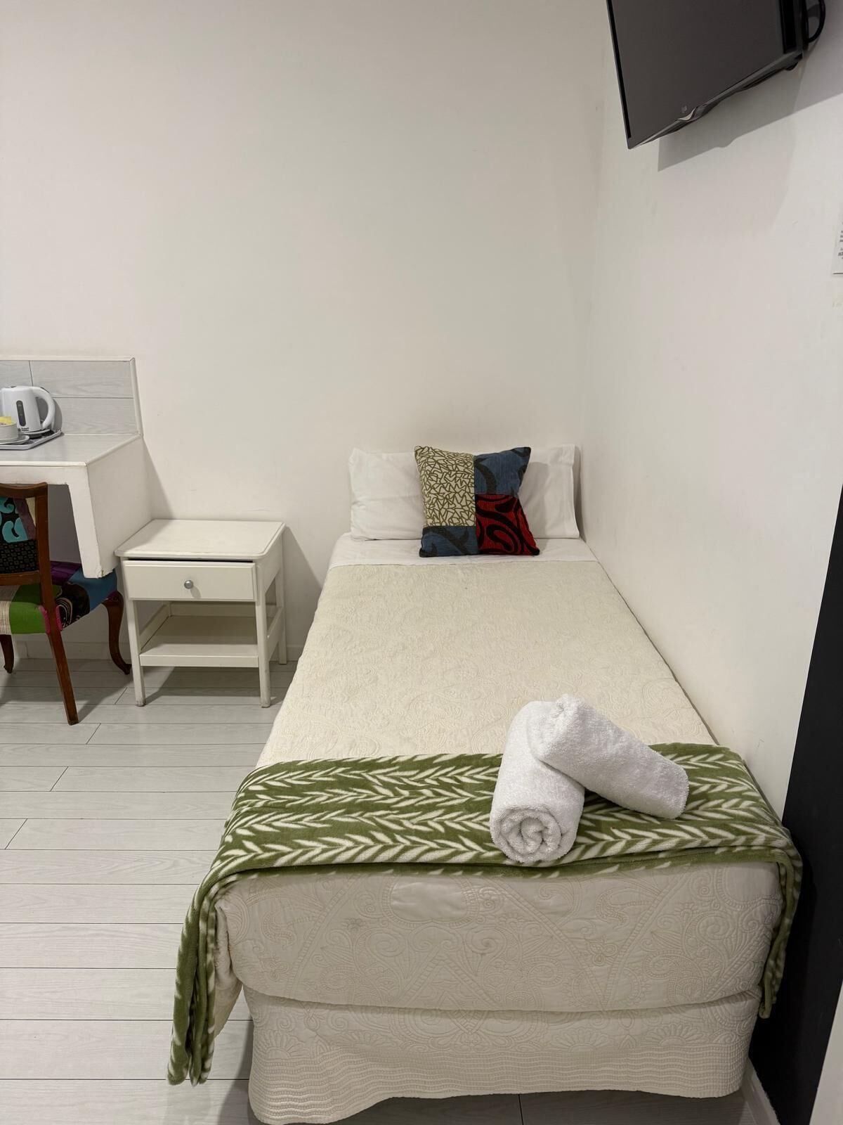 A single bed with a green leaf-patterned blanket, a rolled towel, and two pillows. A white nightstand with a kettle and a small table is beside the bed. A flat-screen TV is mounted on the wall above the bed.