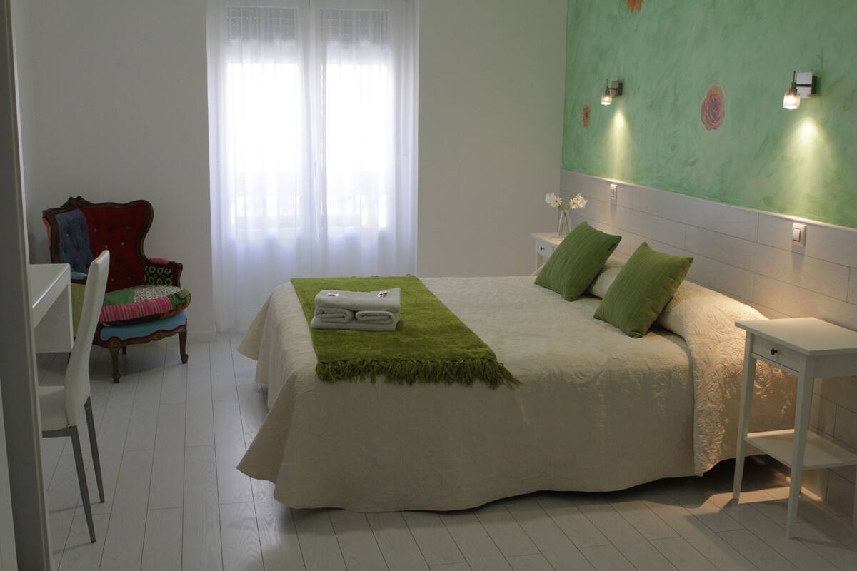 Un dormitorio con cama, una silla, una mesita de noche y una lámpara de pared.