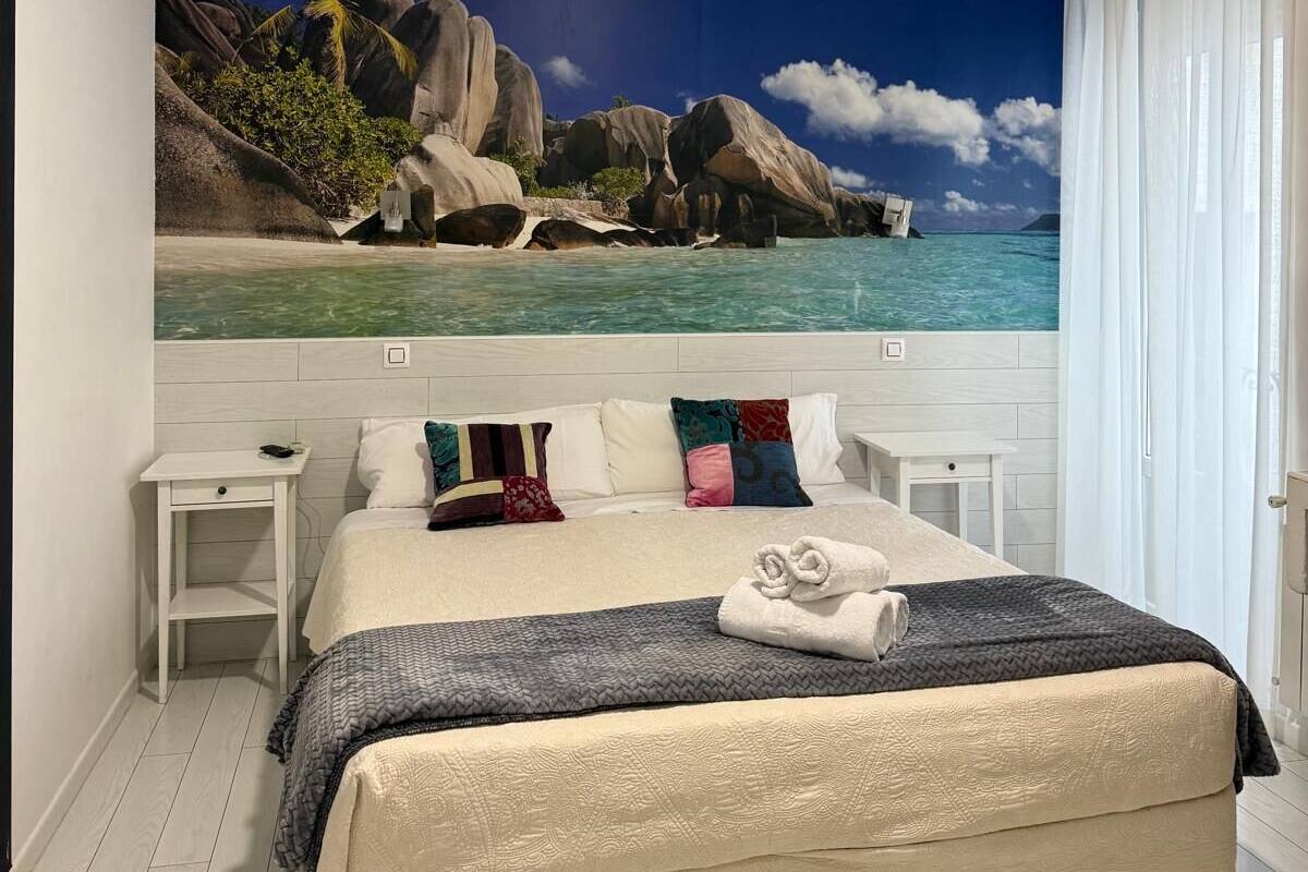 Un dormitorio con una cama grande, una mesita de noche, una pequeña mesa y un mural en la pared con una escena de playa.