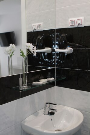 A modern bathroom with a white sink, black tiled walls, and a glass shelf holding a vase with white flowers.