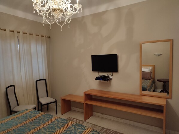 A bedroom with a bed, a TV mounted on the wall, a wooden bench, a chair, and a chandelier. - Villa Ambre (La Marsa)