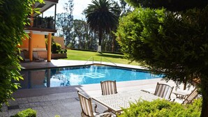 Zona de piscina con sillones de descanso y una mesa, rodeada de vegetación y un edificio con balcón.