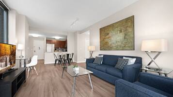 A modern living room with a blue sofa, glass coffee table, and white chairs. A kitchen is visible in the background.