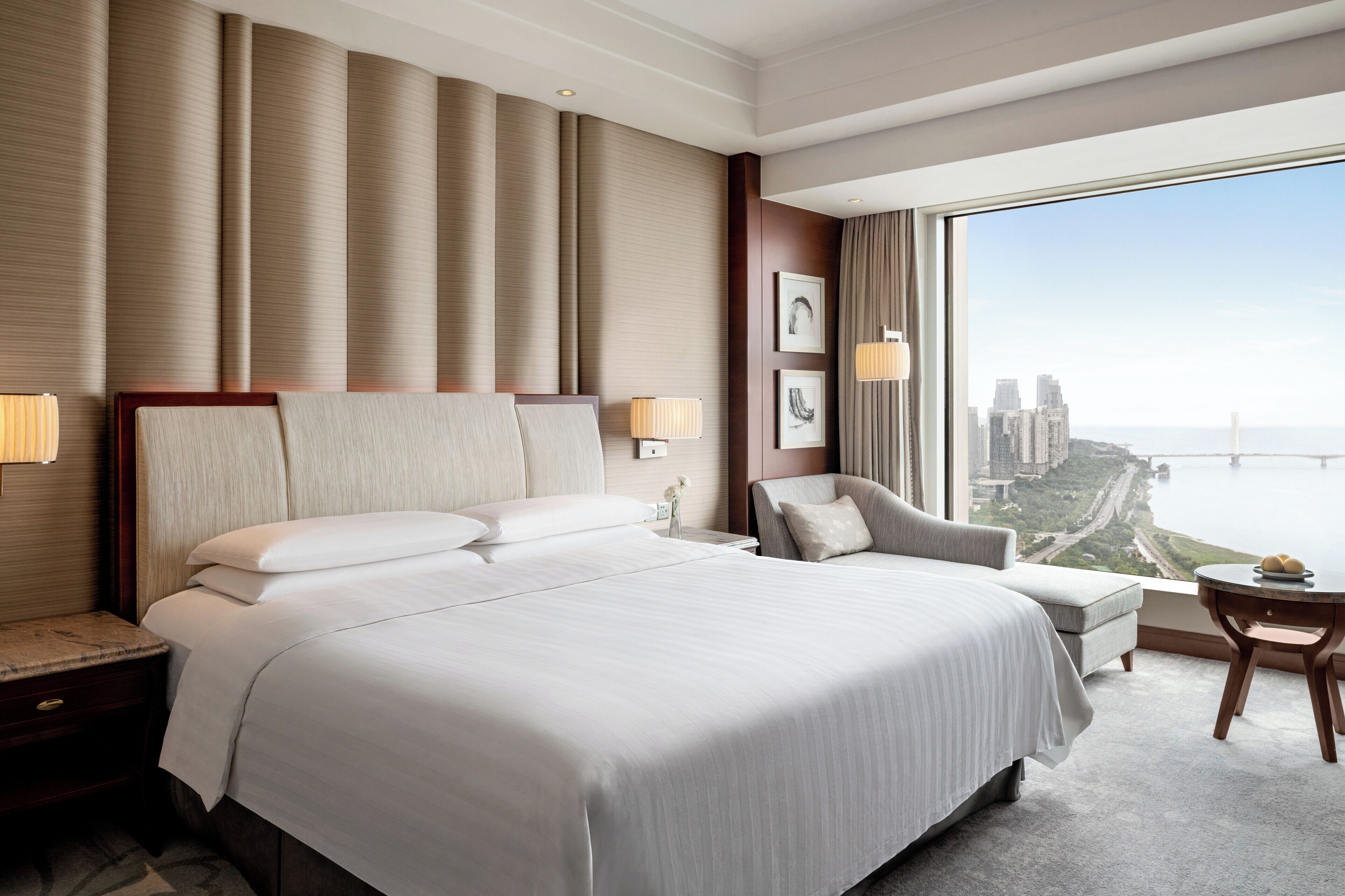 Une chambre d’hôtel moderne, dotée d’un grand lit, d’un bureau, d’une chaise et offrant une vue sur le paysage urbain grâce à une grande fenêtre.