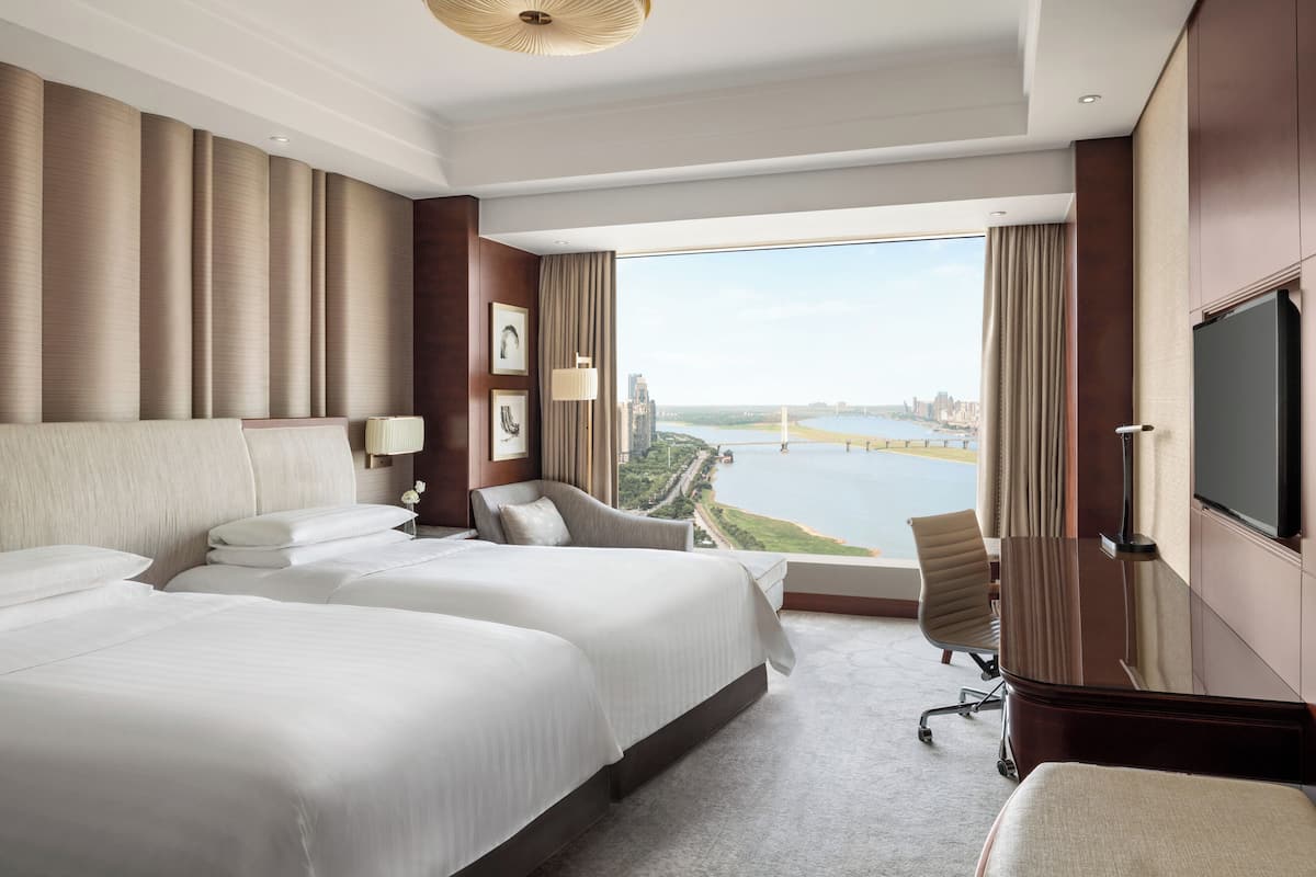 A hotel room with two beds, a TV, a desk, and a view of a river and cityscape.