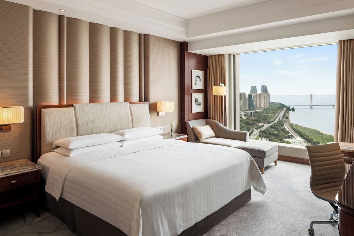 A hotel room with a large bed, a desk, a chair, and a view of a cityscape and bridge.
