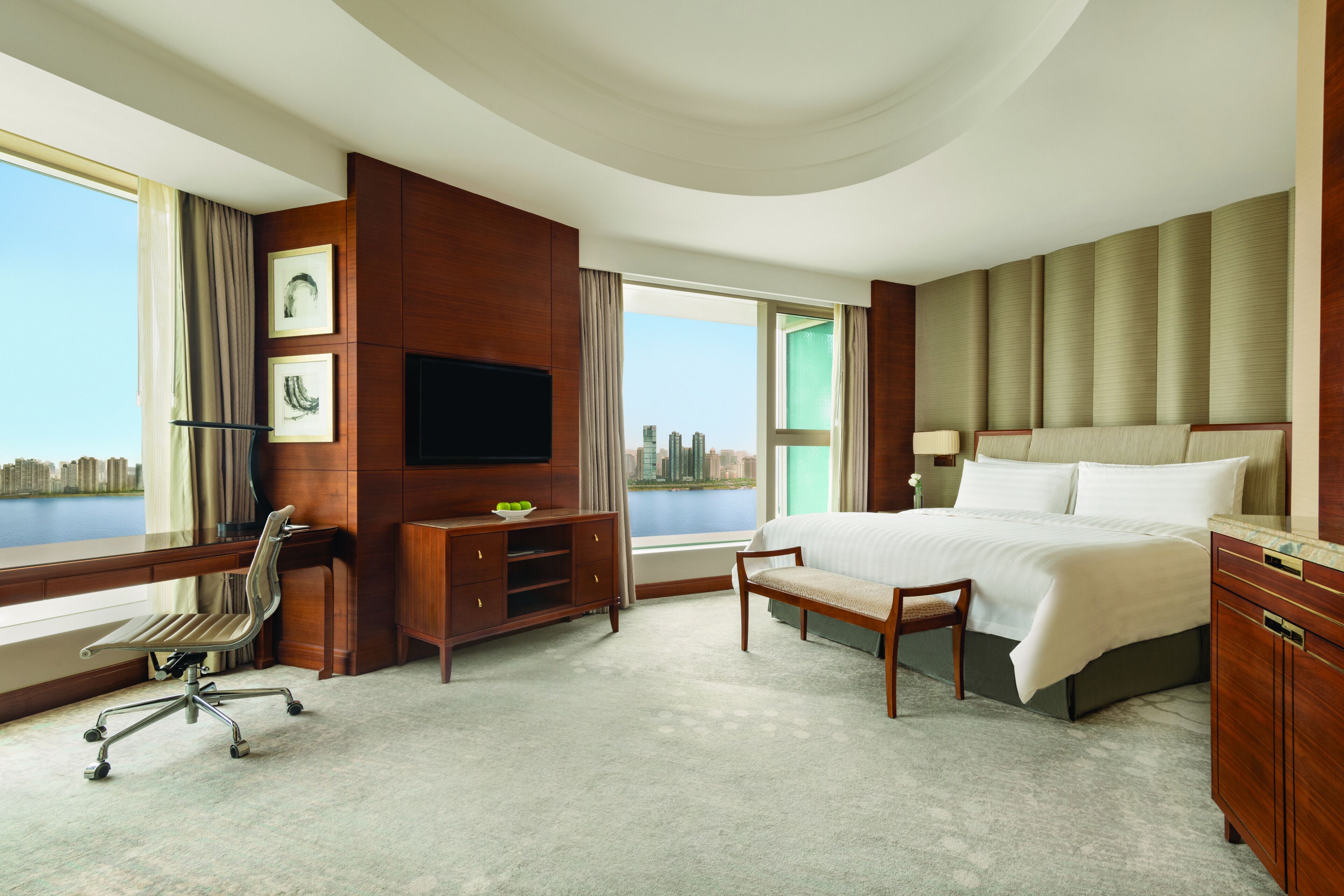 A hotel room with a large bed, a desk, a chair, a TV, and a view of the city.
