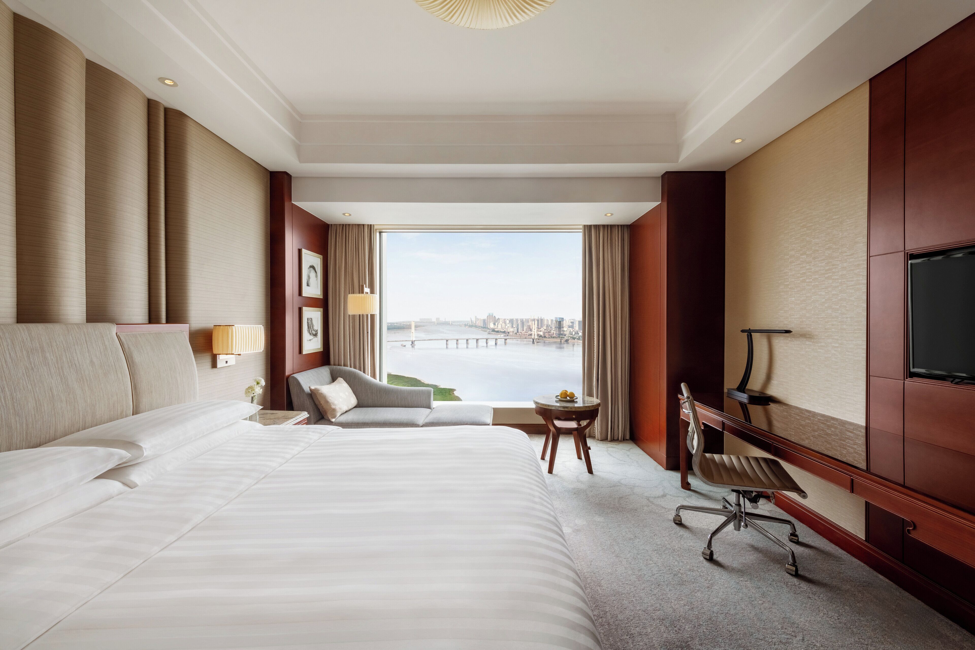 A hotel room with a large bed, a desk, a chair, a TV, and a view of a cityscape.