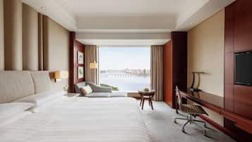 A hotel room with a large bed, a desk, a chair, a TV, and a view of a cityscape.