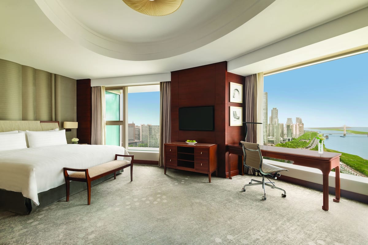 A hotel room with a large bed, a desk, a chair, a TV, and a view of a cityscape and water.