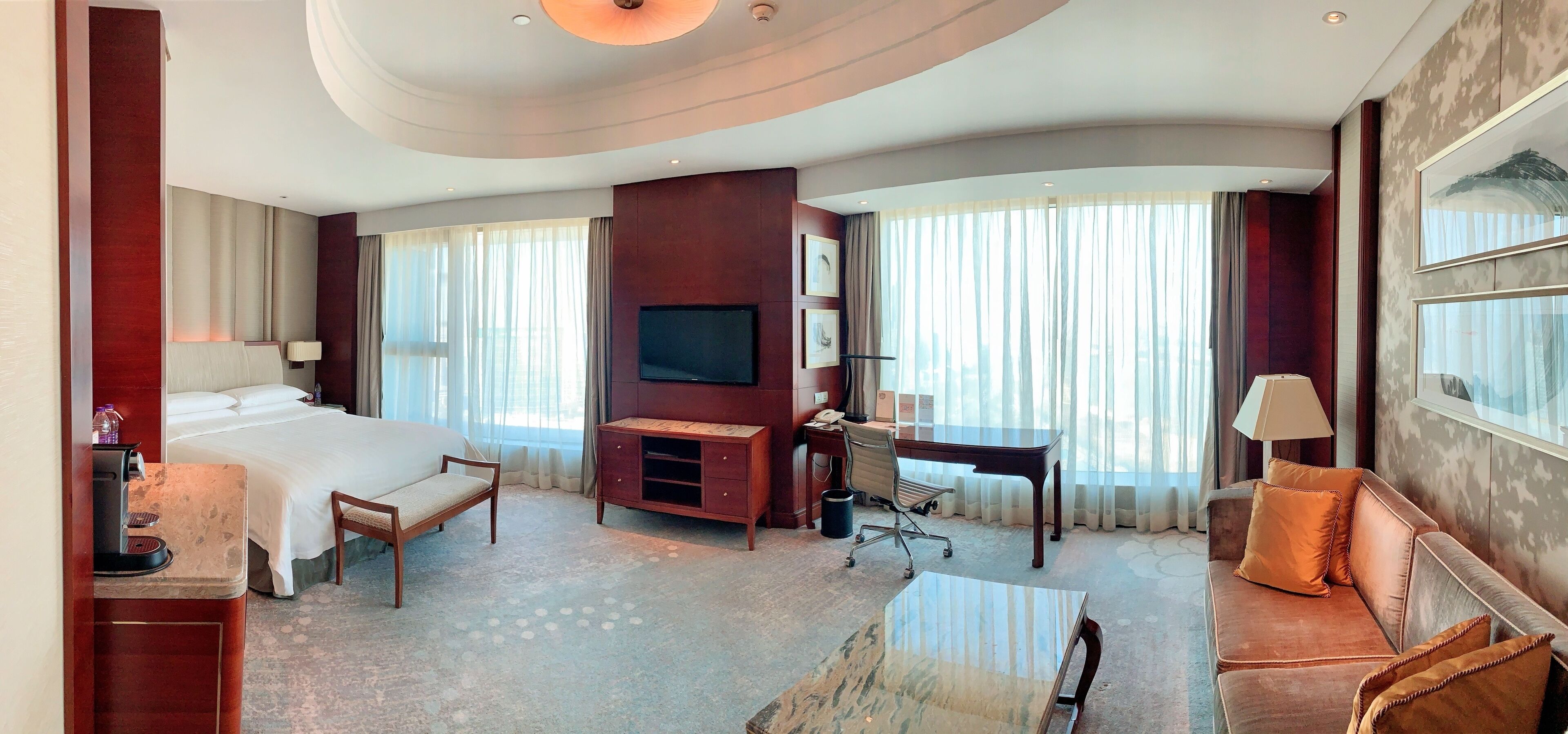 A hotel room with a bed, desk, sofa, TV, and large windows.