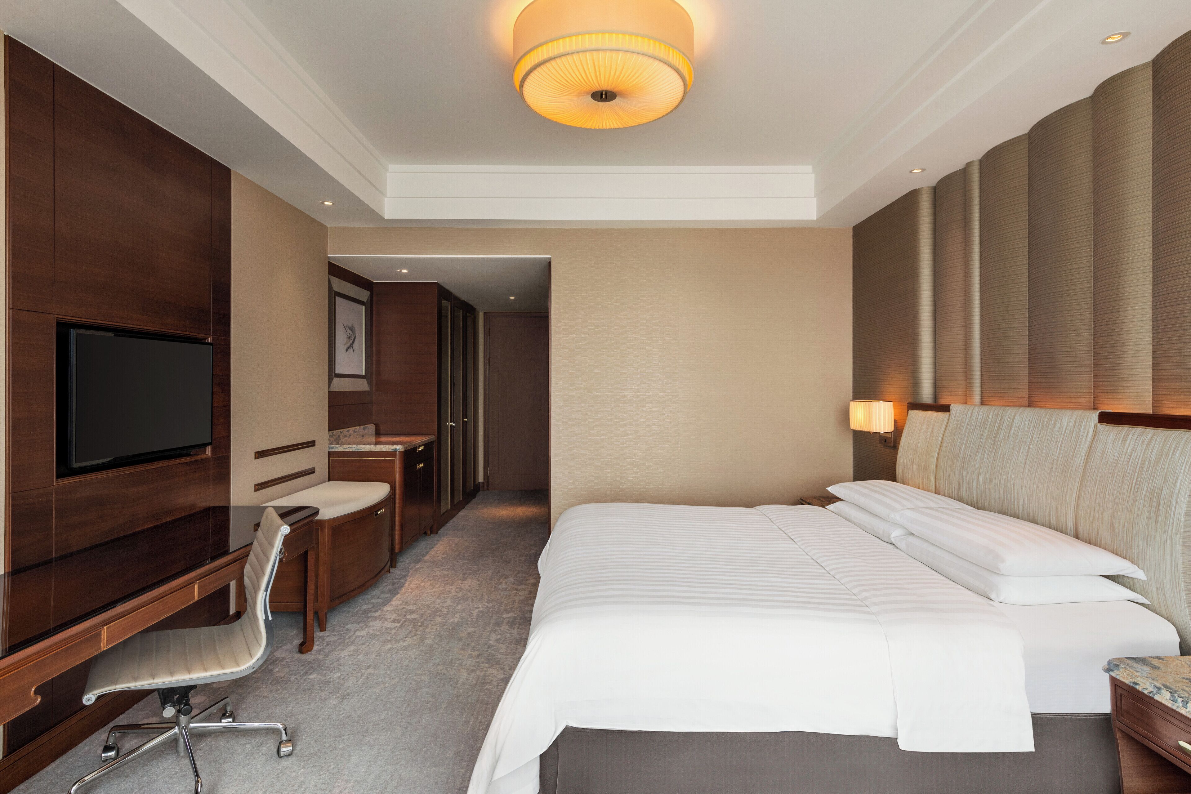A hotel room with a large bed, a desk, a chair, a television, and a wall-mounted lamp.