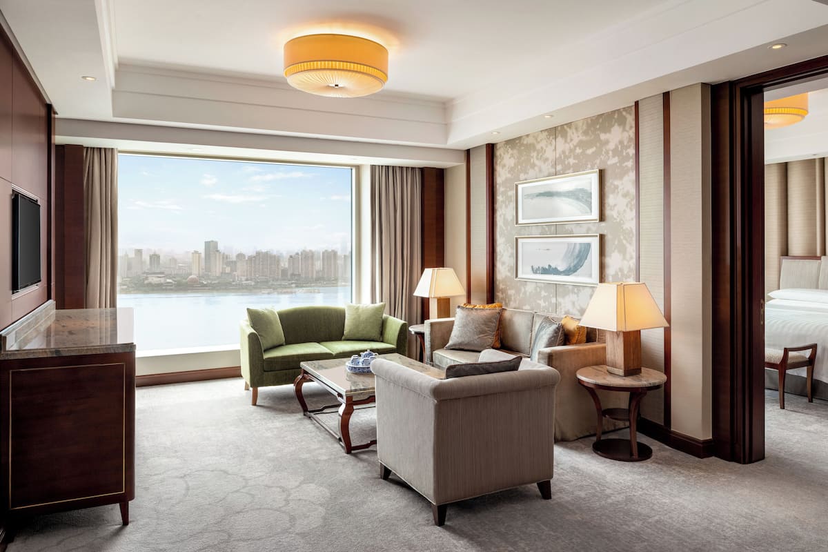 A hotel room with a large window, a green sofa, a beige armchair, a wooden desk, and a bed.