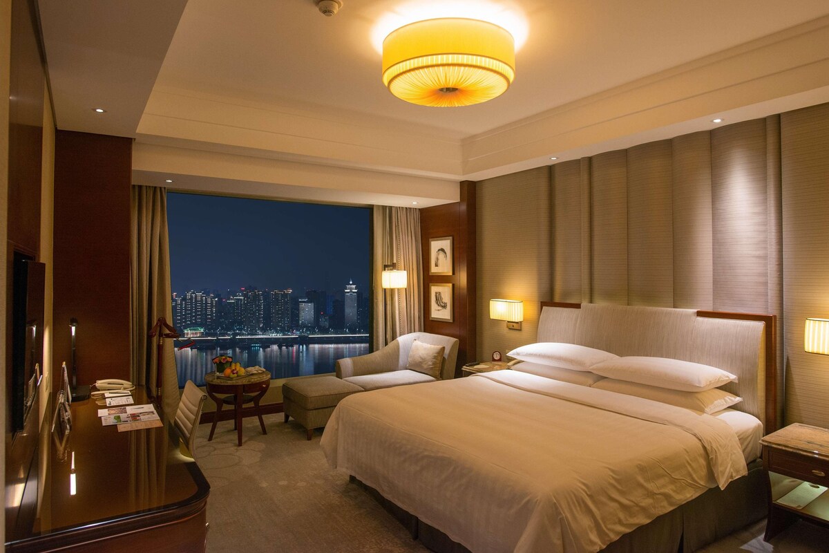 Habitación de hotel con una cama grande, un escritorio, una silla y vistas a un paisaje urbano nocturno.