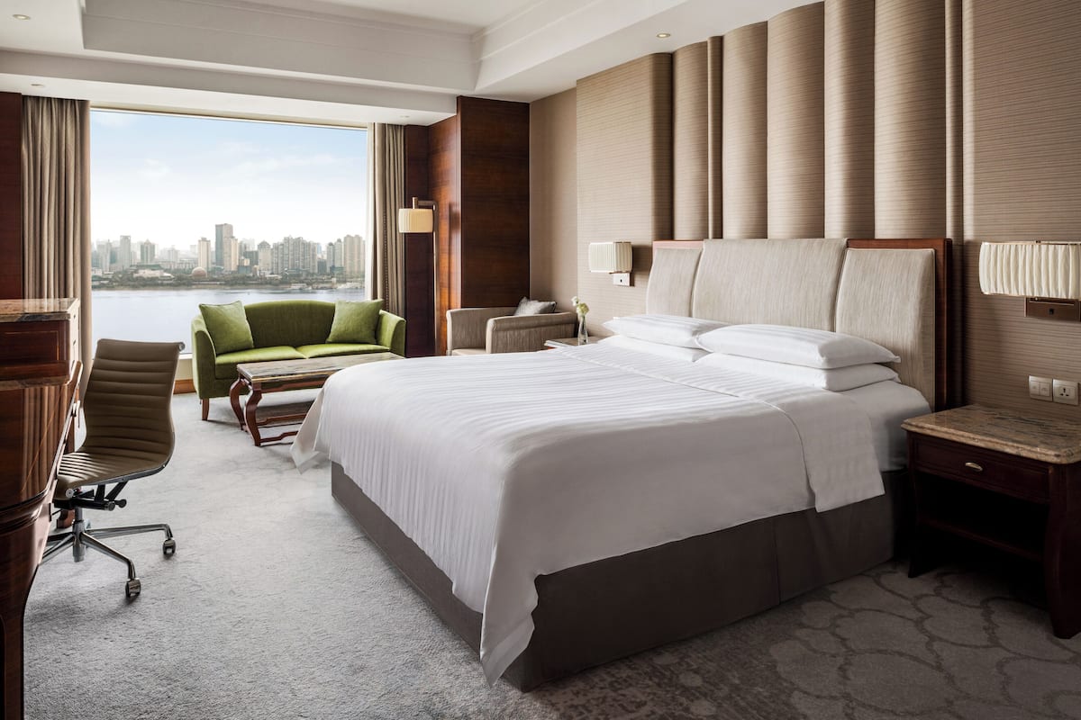 A hotel room with a large bed, a green armchair, a desk, and a view of a city skyline.