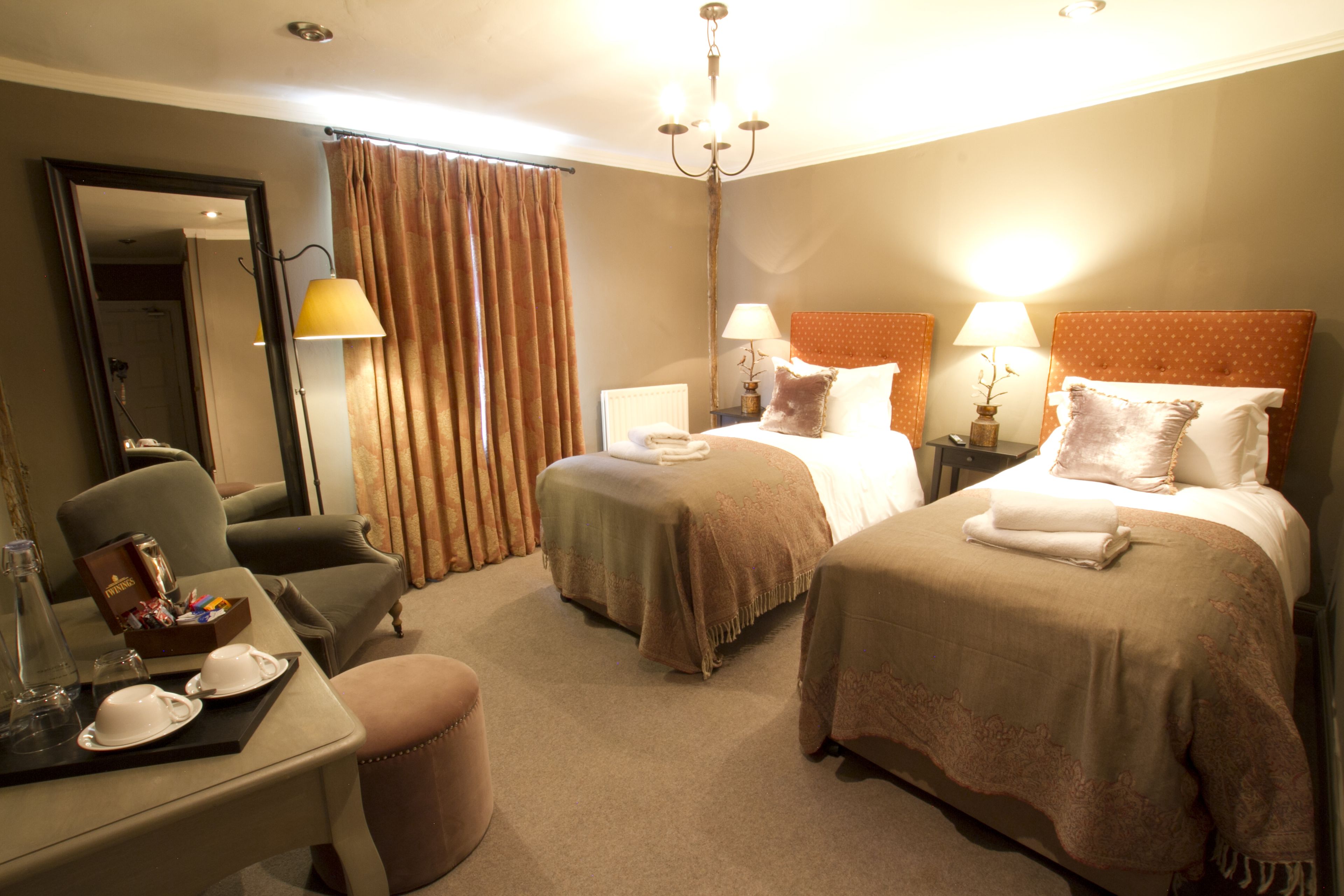 A hotel room with two beds, a sitting area with a chair, a table with a tray of breakfast items, and a mirror.