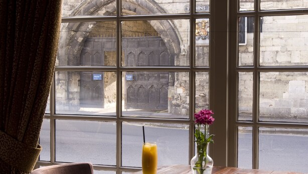 A table with a drink and a vase of flowers, a window with a view of a stone building. - The Chapter House (Salisbury)