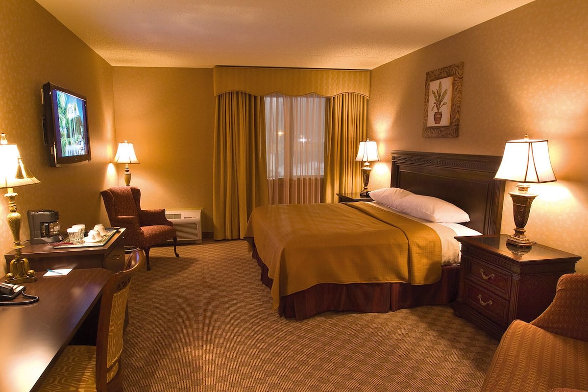Ein Hotelzimmer mit einem Bett, zwei Nachttischen mit Lampen, einem Schreibtisch mit Telefon, einem Sessel, einem Wandtelevisor und einem Fenster mit Vorhängen.