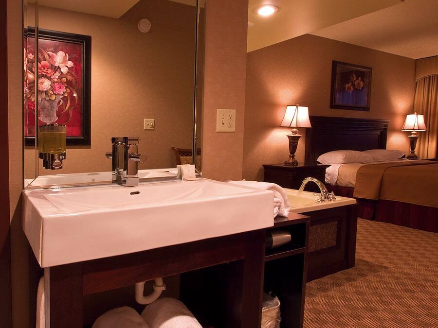 A hotel room with a large mirror, a white sink, a bed, two bedside lamps, and a carpeted floor.