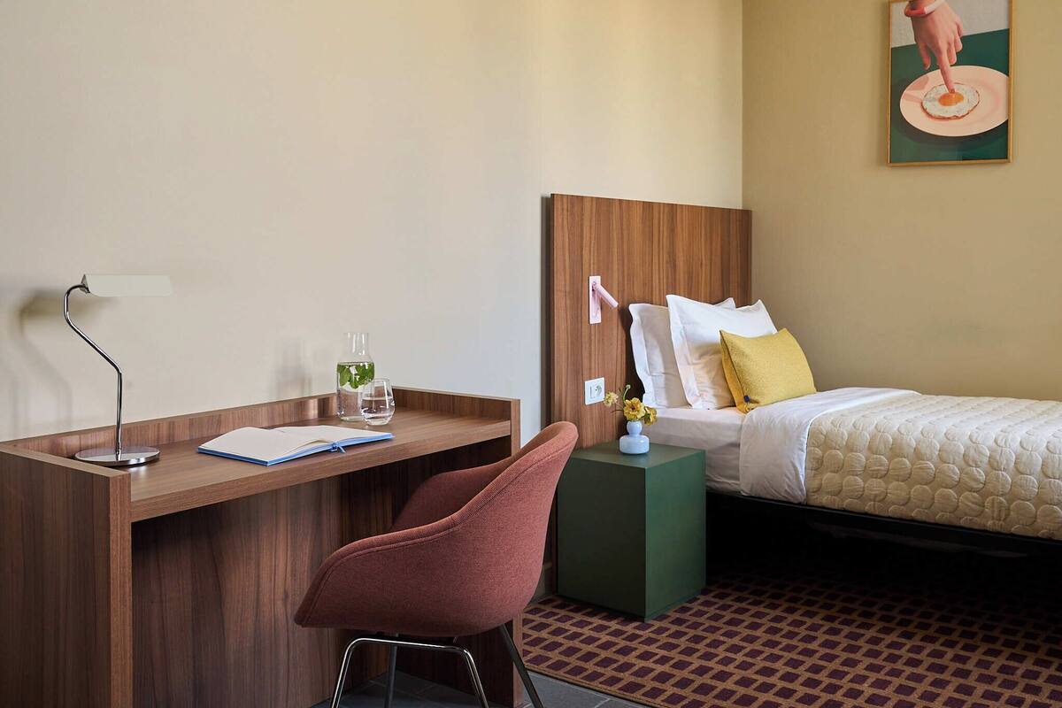 A hotel room with a wooden desk, a chair, a bed with white bedding, and a painting on the wall.