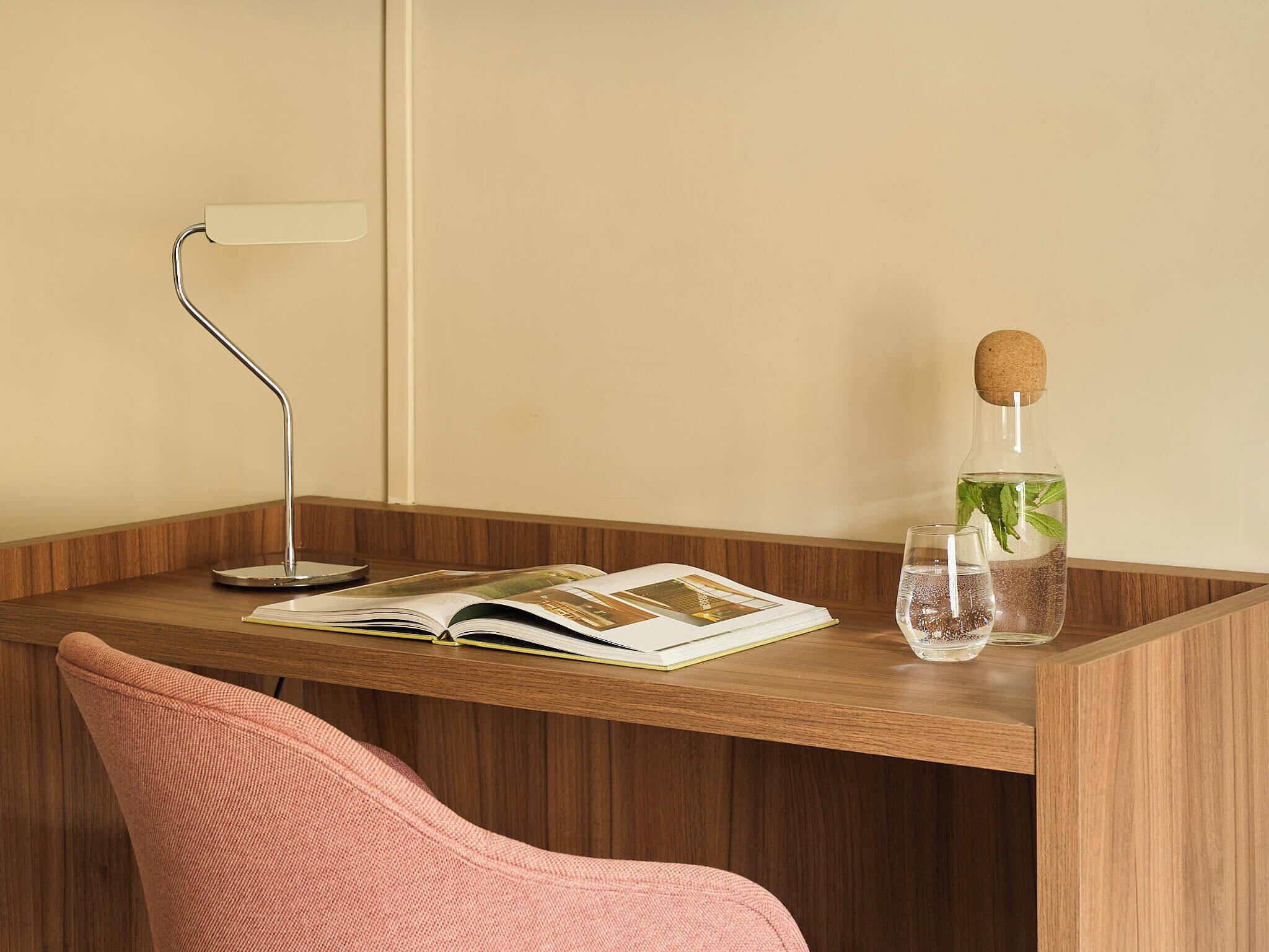 A wooden desk with a book, a glass bottle with a cork stopper, and a metal desk lamp.