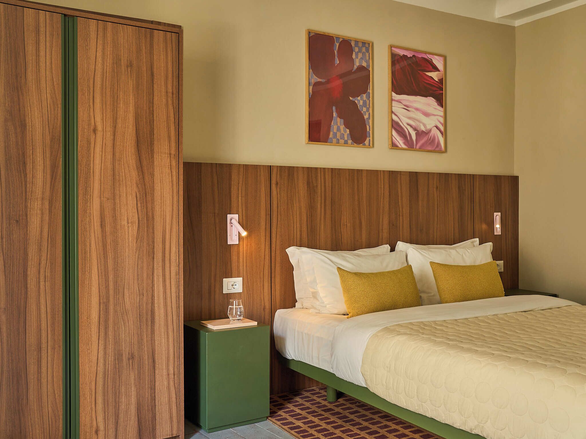 Een slaapkamer met een houten hoofdbord, een bed met witte en mosterdkleurige kussens, een groen nachtkastje en twee abstracte schilderijen aan de muur.