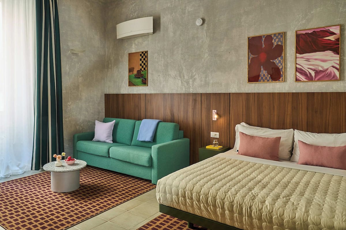 A hotel room with a green sofa, a bed with a quilted duvet, a round coffee table with flowers, and a patterned rug.