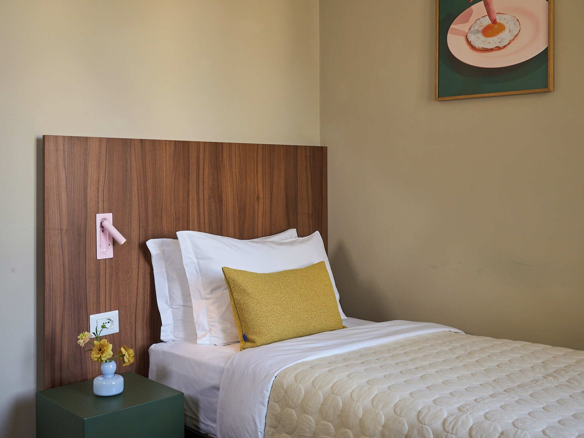 A neatly made bed with white and mustard pillows, a wooden headboard, and a green nightstand with a vase of flowers.