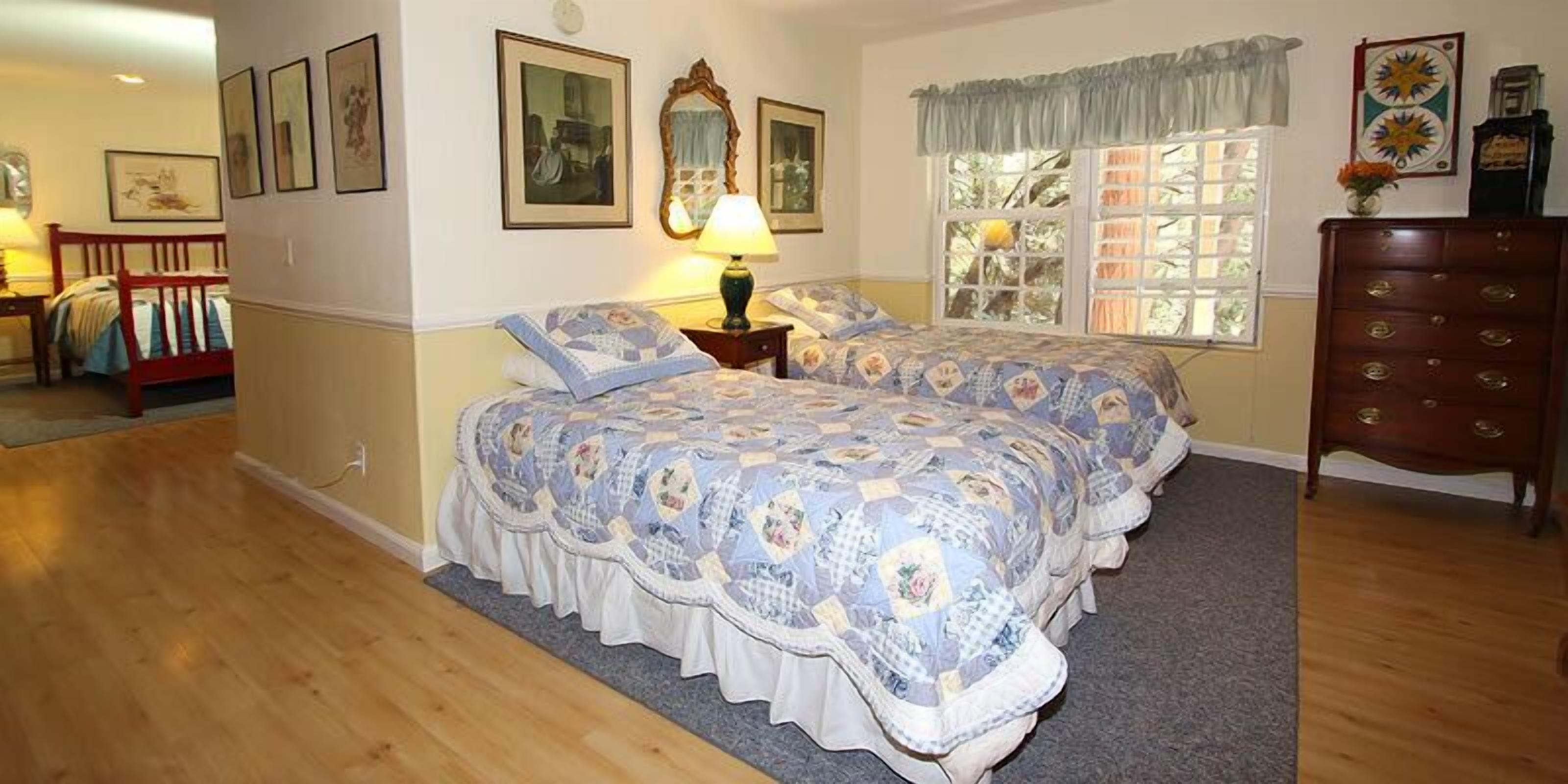Une chambre avec deux lits, une commode, une fenêtre avec des rideaux et des tableaux encadrés accrochés au mur.