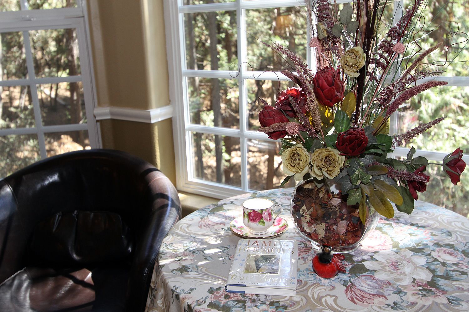 Una mesa con mantel floral, un florero con flores secas, un libro y una taza.