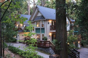 Un chalet à deux étages, avec des fondations en pierre, une terrasse en bois et un grand arbre au premier plan.