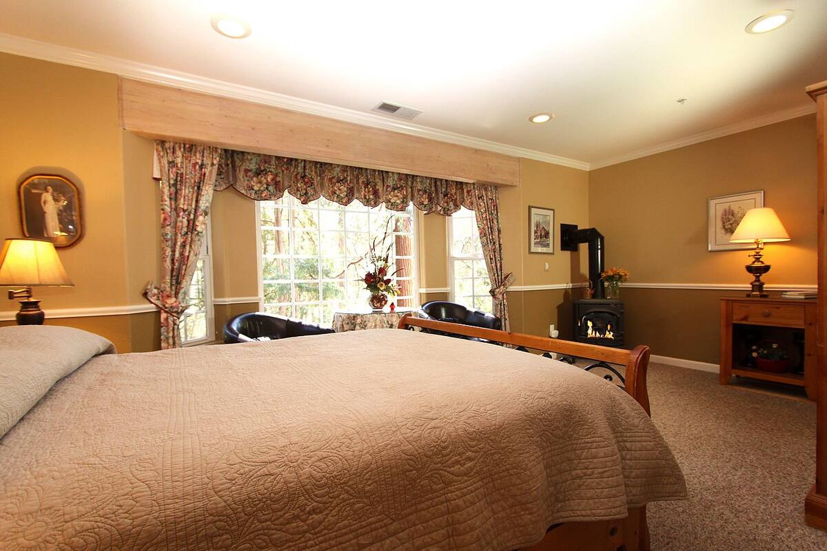 A bedroom with a large bed, a chair, a wooden bench, a fireplace, and a window with curtains.