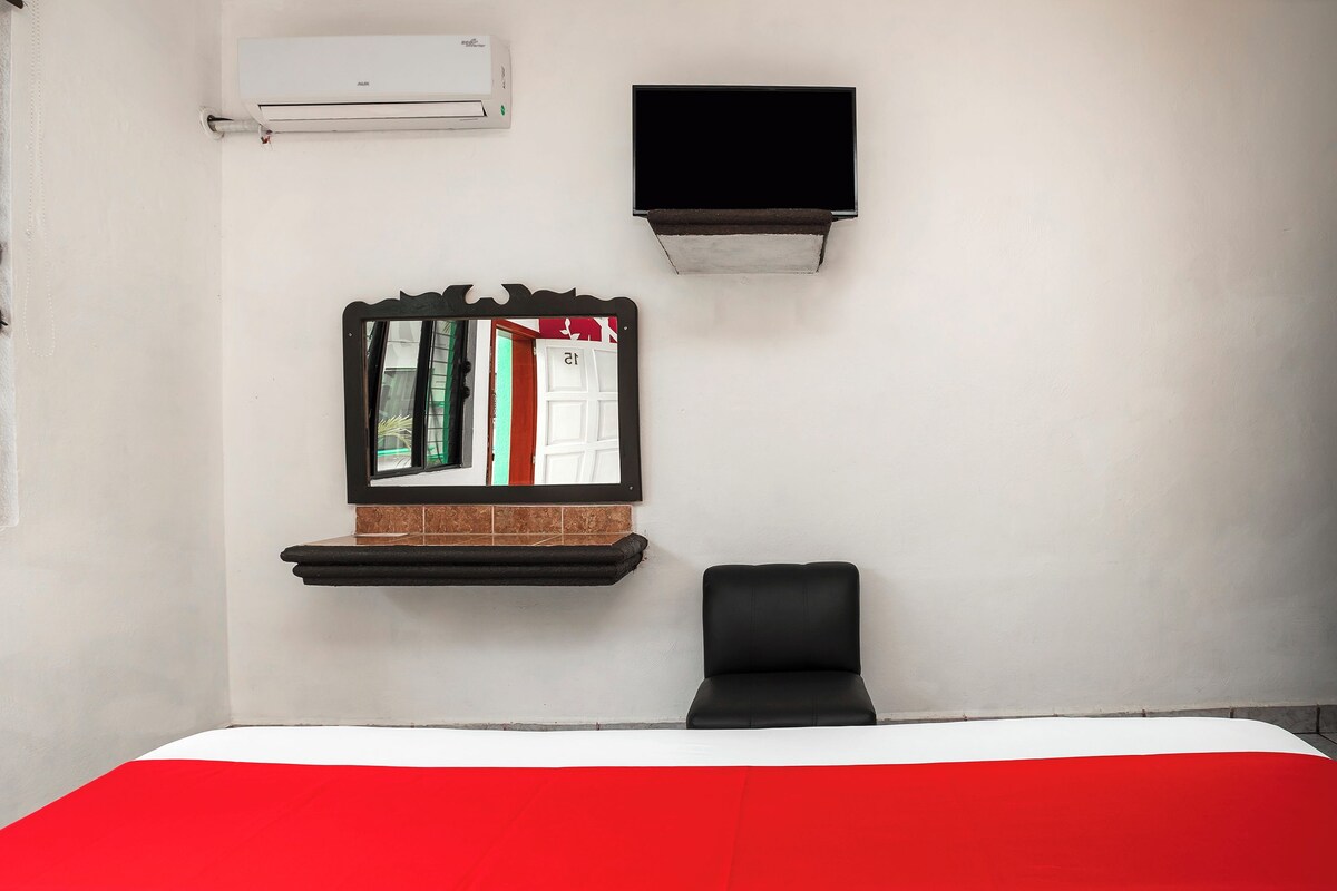 Une chambre d’hôtel comprenant un lit, une chaise, un téléviseur fixé au mur, un climatiseur mural et un miroir avec un cadre décoratif.