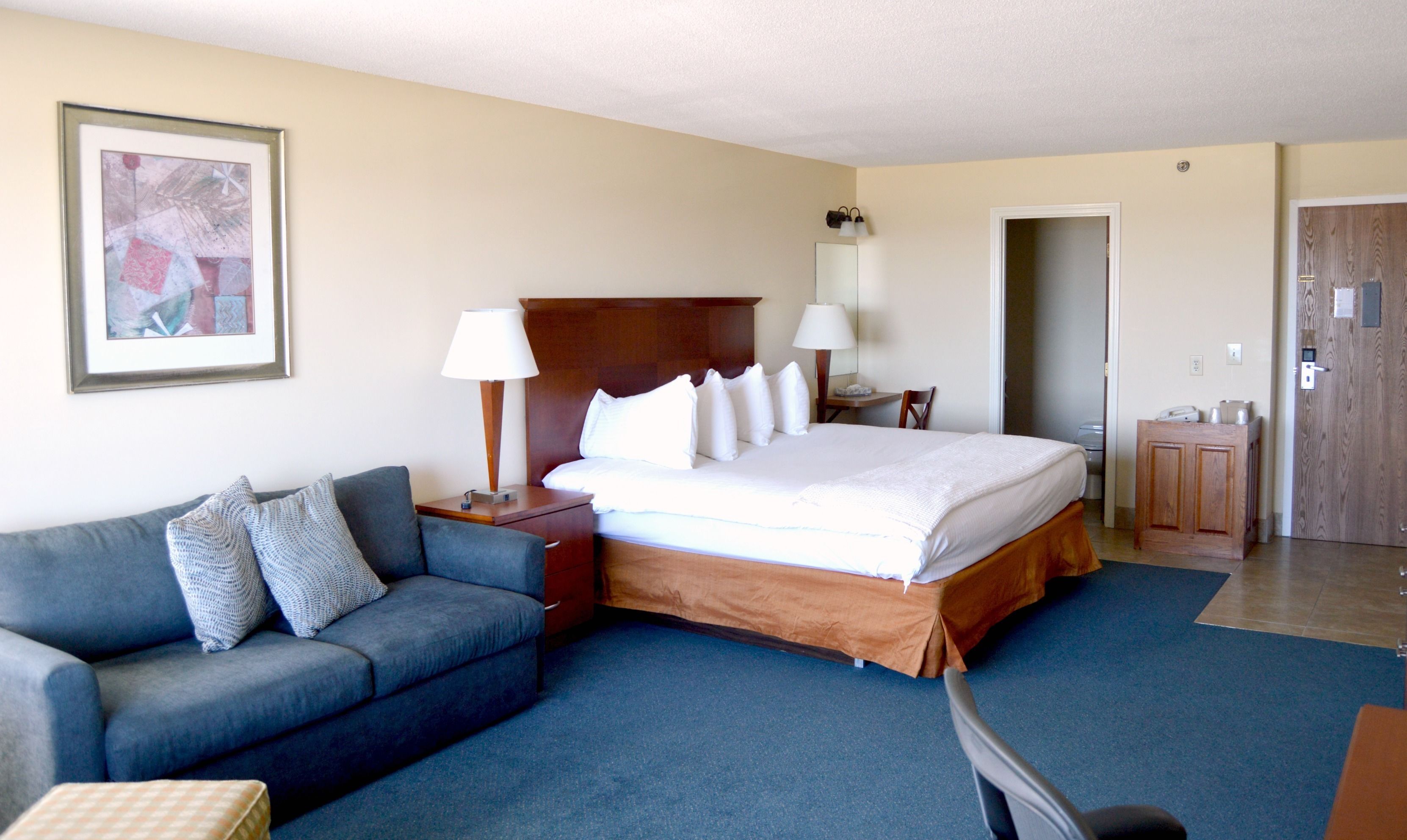 Una habitación de hotel con una cama grande, un sofá, dos mesitas de noche con lámparas, un cuadro enmarcado en la pared y una puerta que da a otra habitación.
