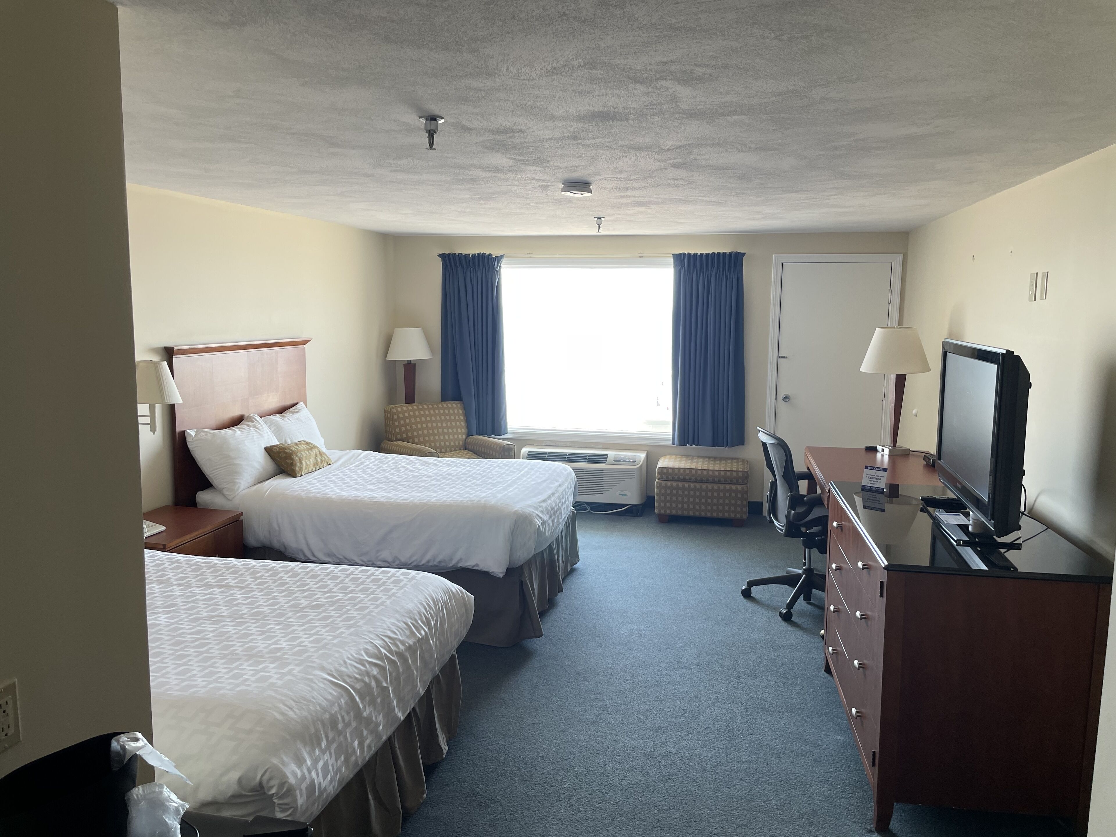 Habitación de hotel con dos camas, un escritorio con televisor, una silla, una lámpara y una ventana con cortinas.