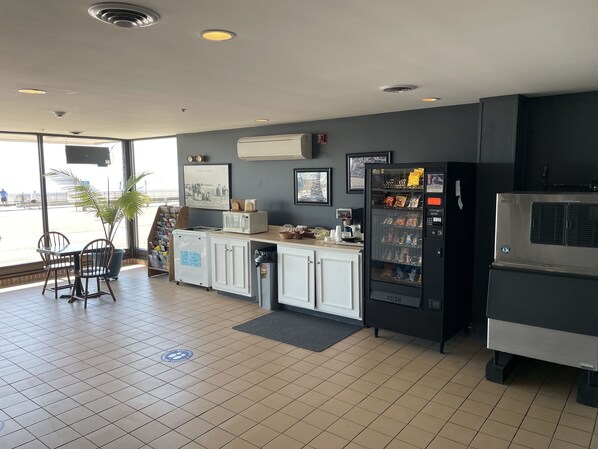 A modern kitchen with a refrigerator, microwave, and coffee maker. There are chairs and a table, and a vending machine with snacks. - Hampton House Hotel (Hampton)
