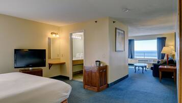A hotel room with a bed, a TV, a desk with a lamp, a chair, a small table, a mirror, and a window with curtains.