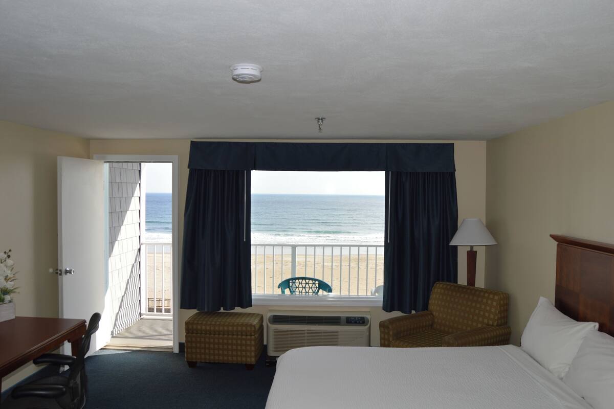 Une chambre d’hôtel dotée d’une grande fenêtre donnant sur la plage, d’un lit, d’un bureau et de deux chaises.
