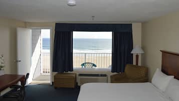 A hotel room with a large window offering a view of the beach, a bed, a desk, and two chairs.