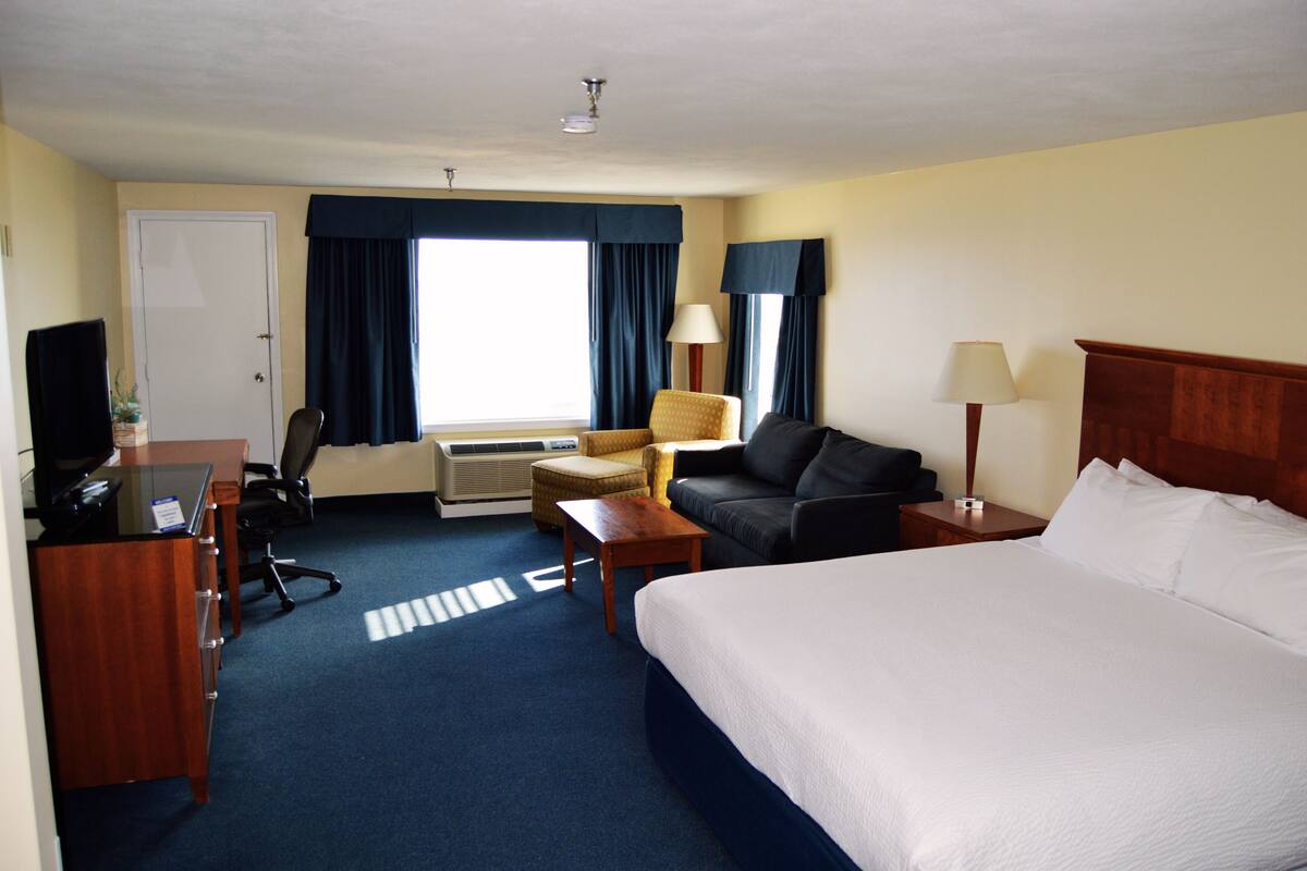 Une chambre d’hôtel comprenant un lit, un bureau avec un ordinateur, une chaise, un canapé, une lampe et une fenêtre avec des rideaux.