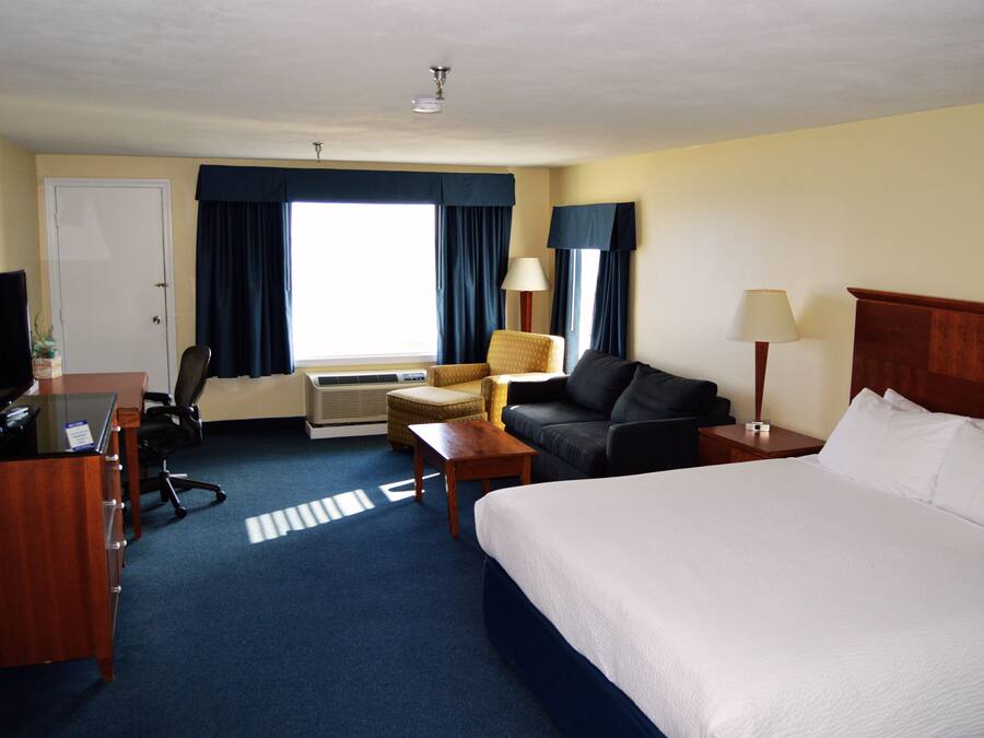 Una habitación de hotel con una cama, un escritorio con una computadora, una silla, un sofá, una lámpara y una ventana con cortinas.