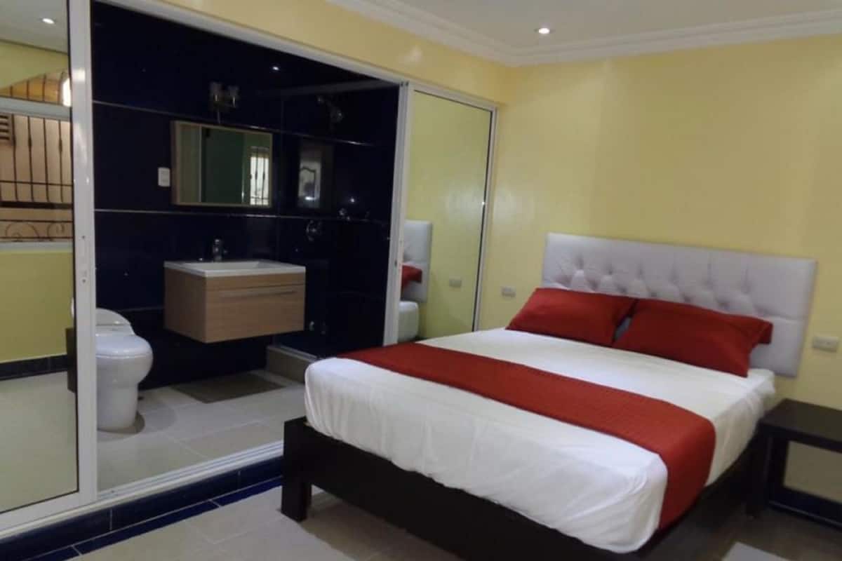 A bedroom with a bed, a nightstand, a mirror, a toilet, and a bathroom vanity.